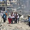 Health workers and a person in a white helmet carry an injured person on a stretcher through a war-torn street filled with rubble and debris.