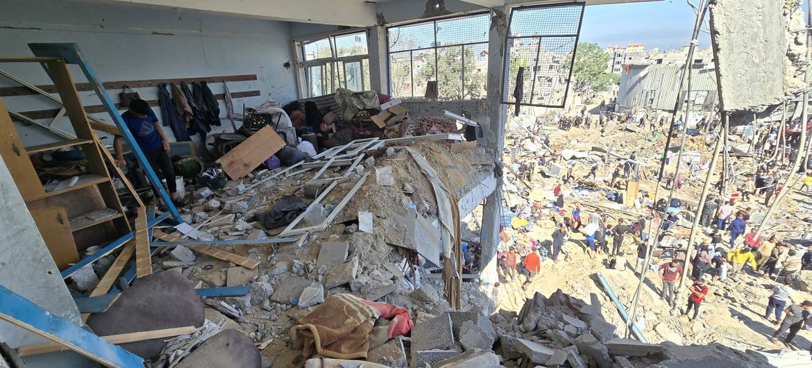 View from inside a damaged UNRWA shelter in Al Bureij, Gaza, showing rubble and debris, with displaced families and survivors visible outside.