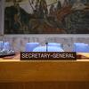 The Secretary-General's nameplate on a wooden desk in the UN Security Council Chamber, with blue chairs and a large mural in the background.