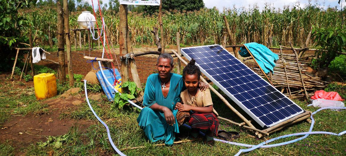 A woman and child sit next to a solar panel and a water pump system in a field in Dhanghista, Ethiopia.