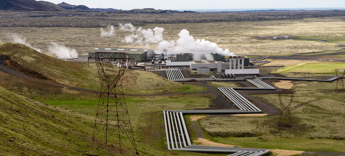 A wide shot of the Hellisheiði Geothermal Power Plant in Iceland, showing steam rising from the facility and large pipes snaking across the green, hilly landscape.