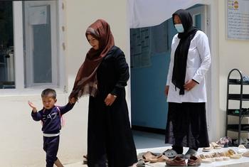 A midwife in a white coat and face mask stands at a clinic entrance in Afghanistan, while a woman in a brown hijab walks a young boy past the door.