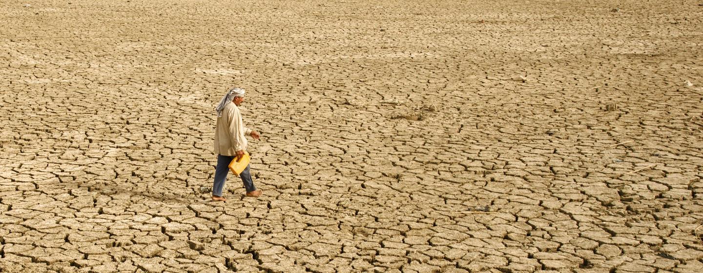 A man walks across a vast, cracked, and dry landscape in Iraq, illustrating the severe impacts of climate change and drought.