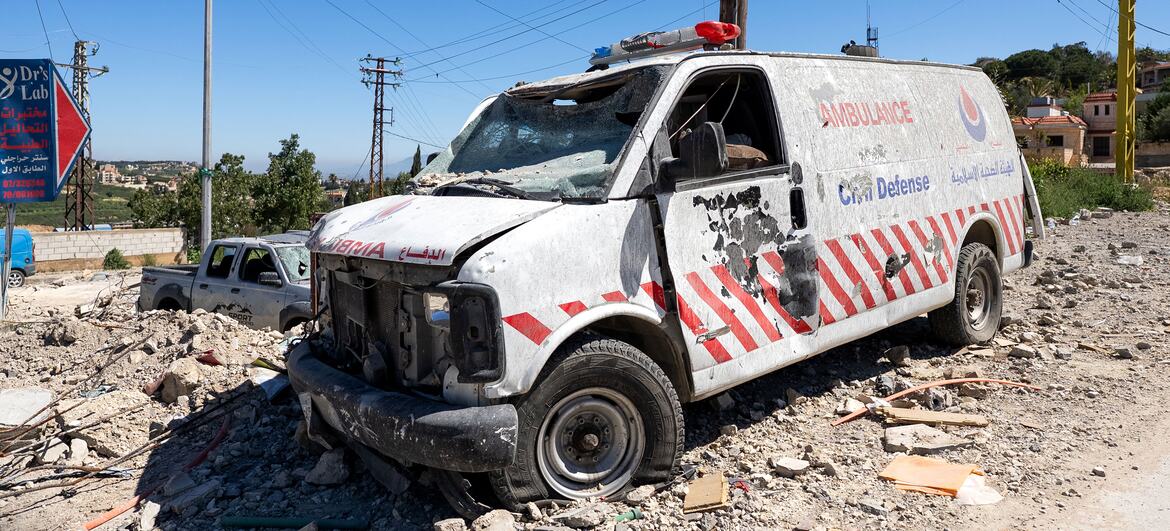 A severely damaged white ambulance with shattered windows and bullet holes sits amidst rubble on a street in Tebnine, Lebanon, showing the impact of conflict on emergency services.