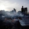Silhouettes of people stand amidst the ruins of a displaced persons camp in Rafah, Gaza, with smoke and fire rising from the wreckage.