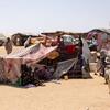 Displaced families live in temporary shelters at the Goz Alsalam IDP site in Sudan. Women and children sit outside a makeshift tent made from patterned fabric, while others walk in the sandy encampment.