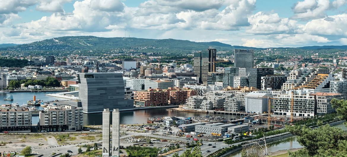 Panoramic view of Oslo, Norway, featuring modern skyscrapers, the harbor, and surrounding hills under a cloudy blue sky.
