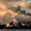 A view of a polluted industrial waterfront at sunset, with large plumes of dark smoke billowing from factory smokestacks into the sky. Several large ships are docked in the foreground on the water.