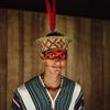 Portrait of a young indigenous man in traditional attire with face paint and a feathered headdress, standing in front of a wooden wall.