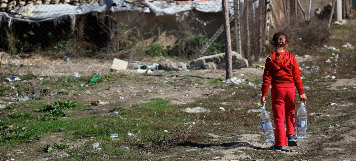 A young girl in red clothing walks away from the camera, carrying two large plastic water bottles. In the background are makeshift structures with corrugated metal and tiled roofs in a rural setting.
