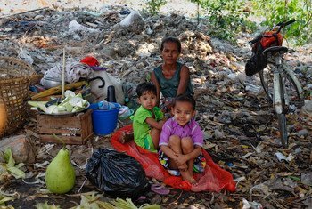 A homeless family, consisting of a woman and two young children, sits on a red mat in an outdoor area filled with garbage and debris in Yangon, Myanmar.