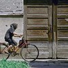 An elderly woman in a polka dot dress and headscarf rides a bicycle past a weathered wooden door.